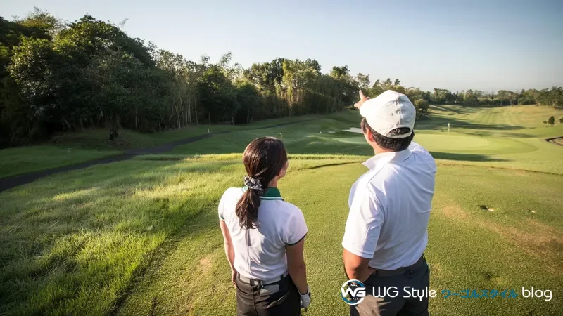 30代の日本人男女がコース高台からホール全景（フェアウェイ・バンカー・グリーン）を眺めている後ろ姿